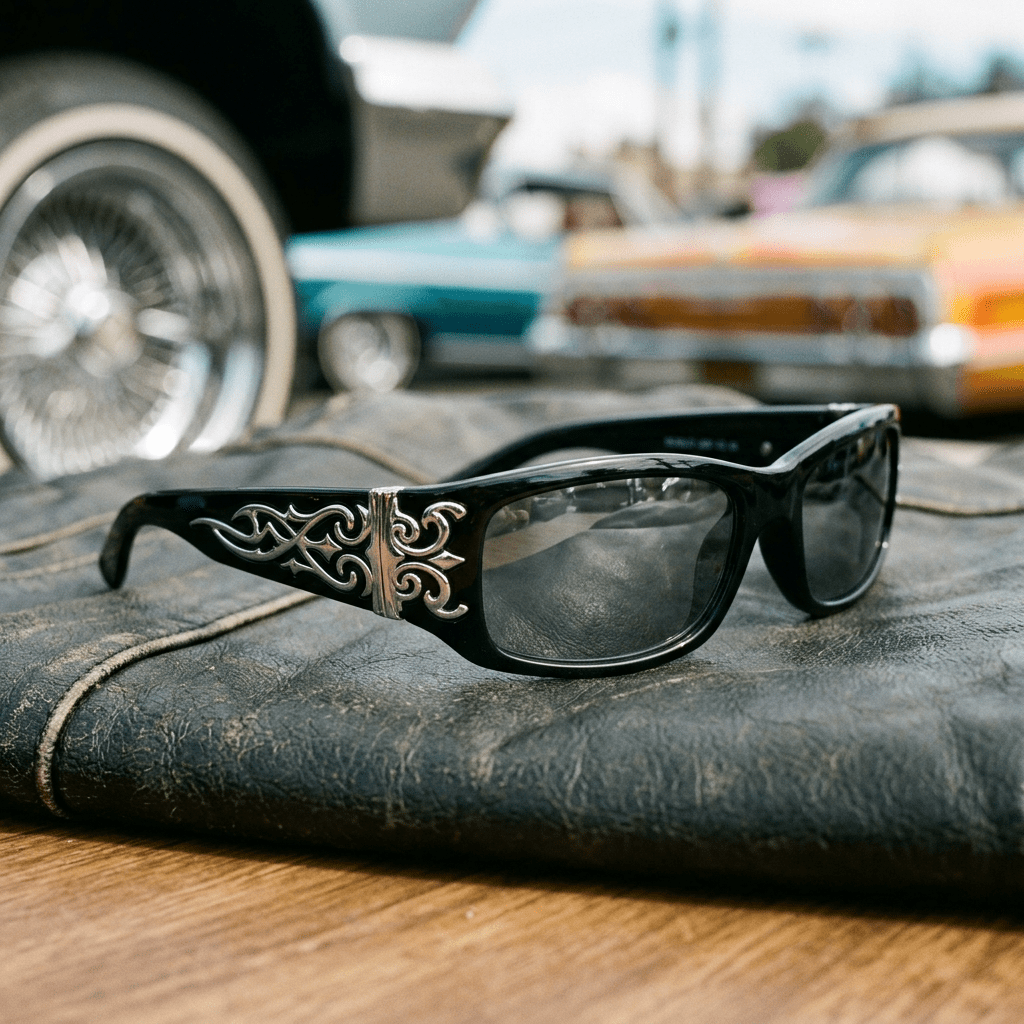Black sunglasses with silver decorative patterns on the temples placed on a worn leather surface with vintage cars blurred in the background